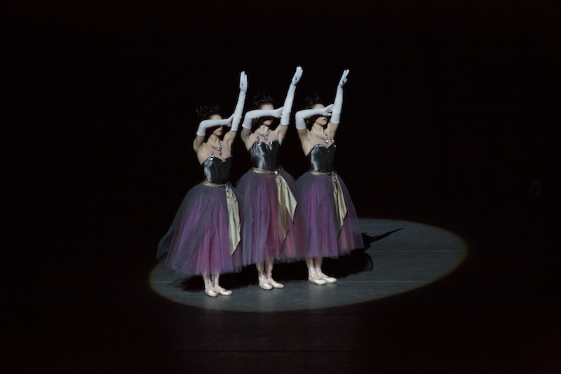 Three dancers in purple tutus and slinky white gloves cover their eyes with their forearm while their other arm extends above them